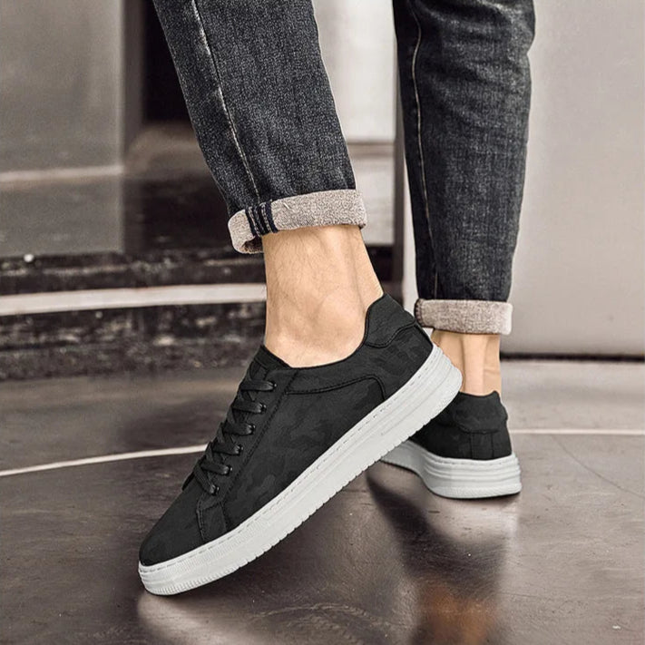 Black sneakers with white soles worn with rolled-up jeans on a reflective floor.