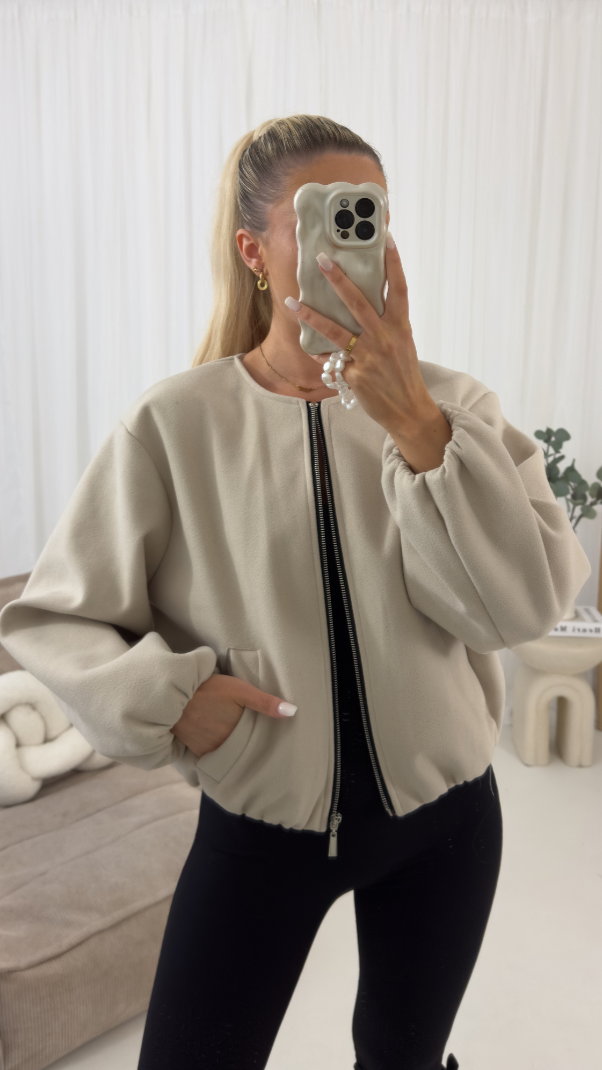 Person wearing a beige bomber jacket taking a mirror selfie in a room with white walls and a couch.
