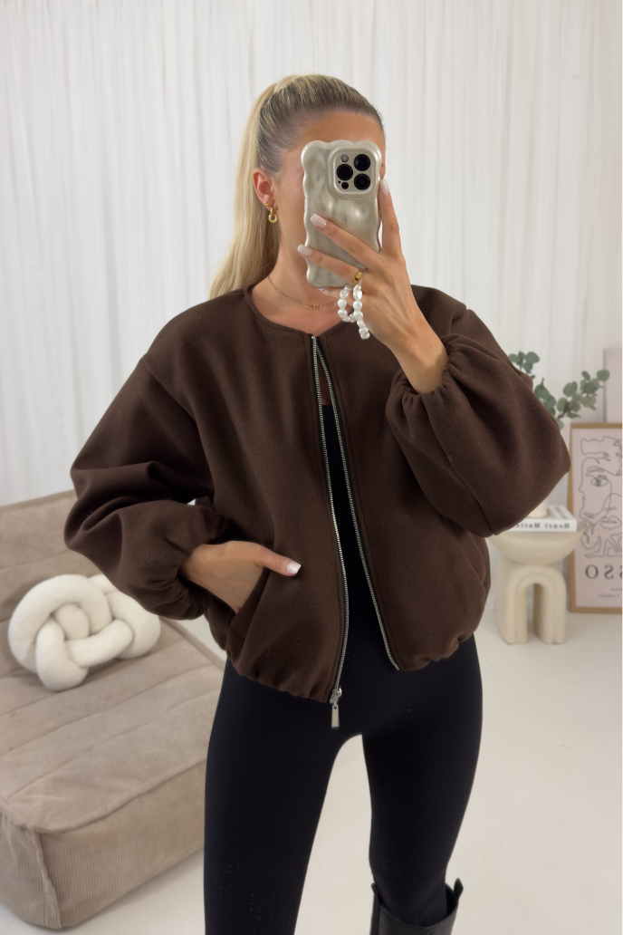 Person wearing a brown bomber jacket in a room with a beige couch and white wall.