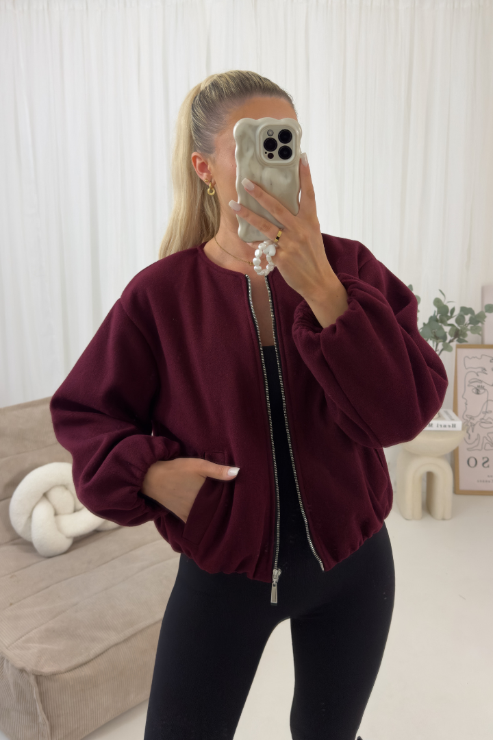 Person wearing a burgundy bomber jacket and black pants in a room with a white curtain and beige sofa.