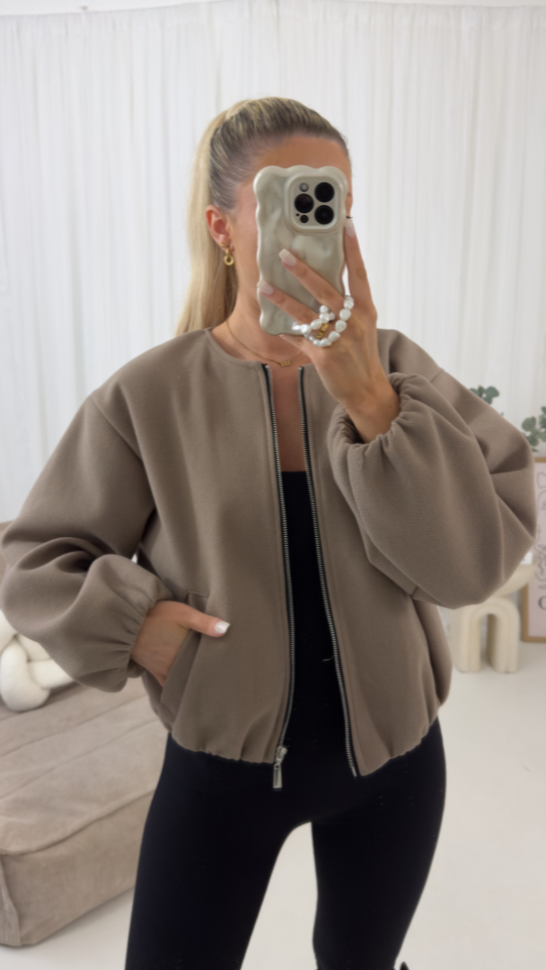 Person wearing a beige jacket taking a mirror selfie in a room with white walls.