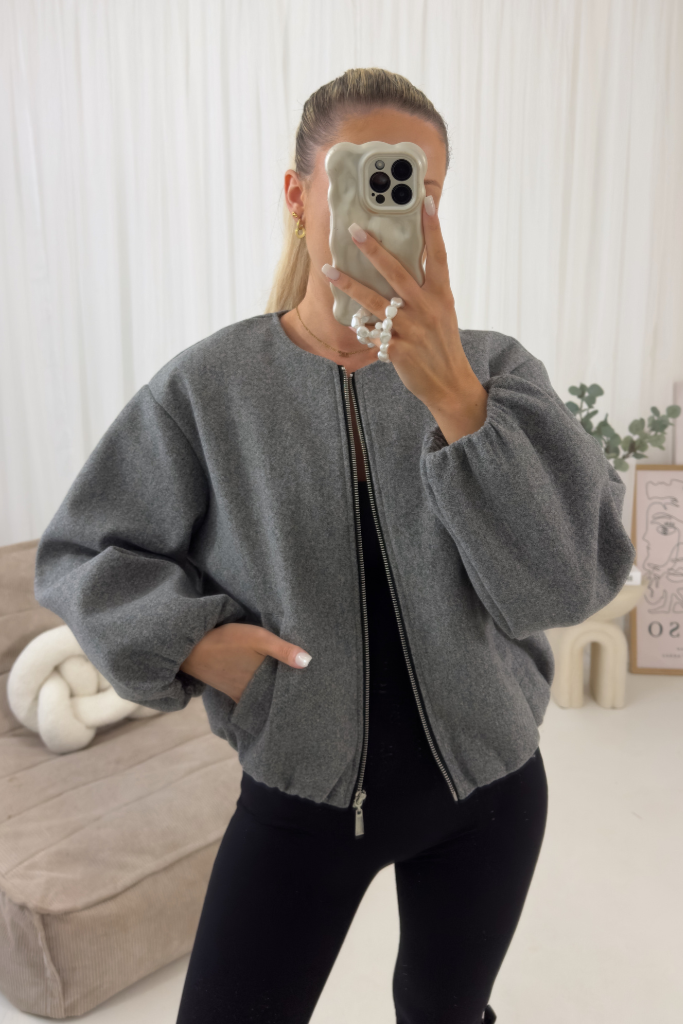 Person wearing a gray jacket taking a mirror selfie in a minimalistic room.