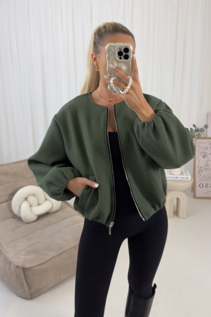 Person wearing a green jacket taking a mirror selfie in a room with a beige couch and decorative items.