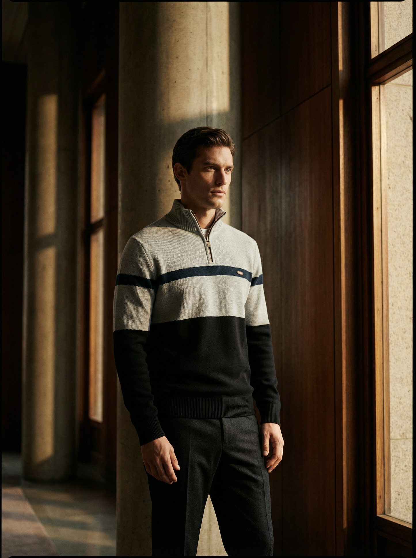Man wearing a striped sweater and dark pants standing in a sunlit hallway.