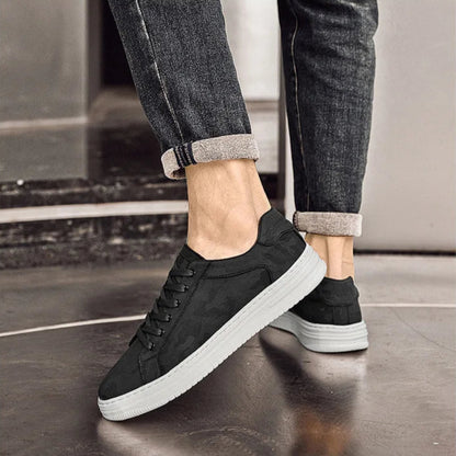Black sneakers with white soles worn with rolled-up jeans on a reflective floor.