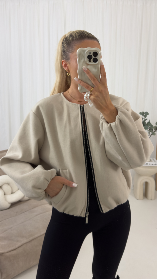 Person wearing a beige bomber jacket taking a mirror selfie in a room with white walls and a couch.