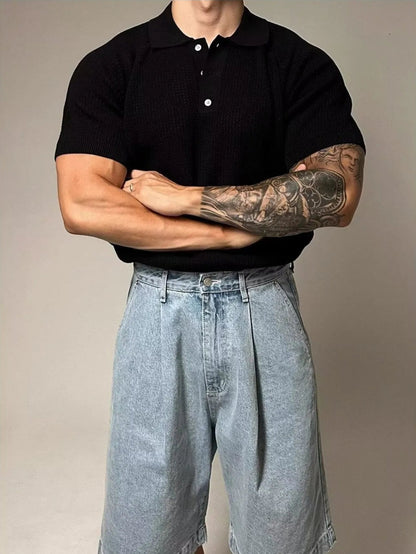 Person wearing a black polo shirt and light blue denim shorts with tattoos on arms.