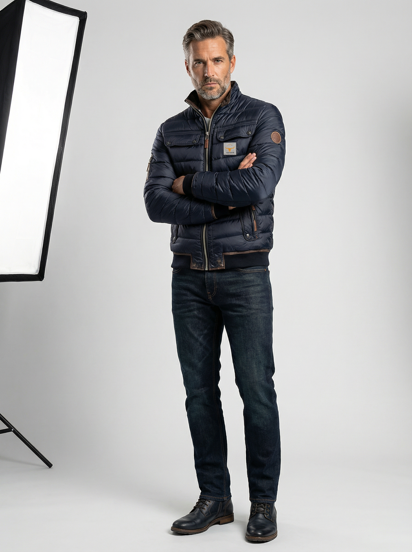 Man wearing a dark jacket and jeans standing in front of a photography light stand on a white background
