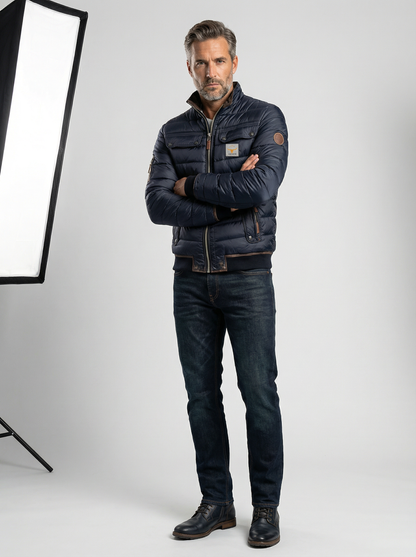 Man wearing a dark jacket and jeans standing in front of a photography light stand on a white background