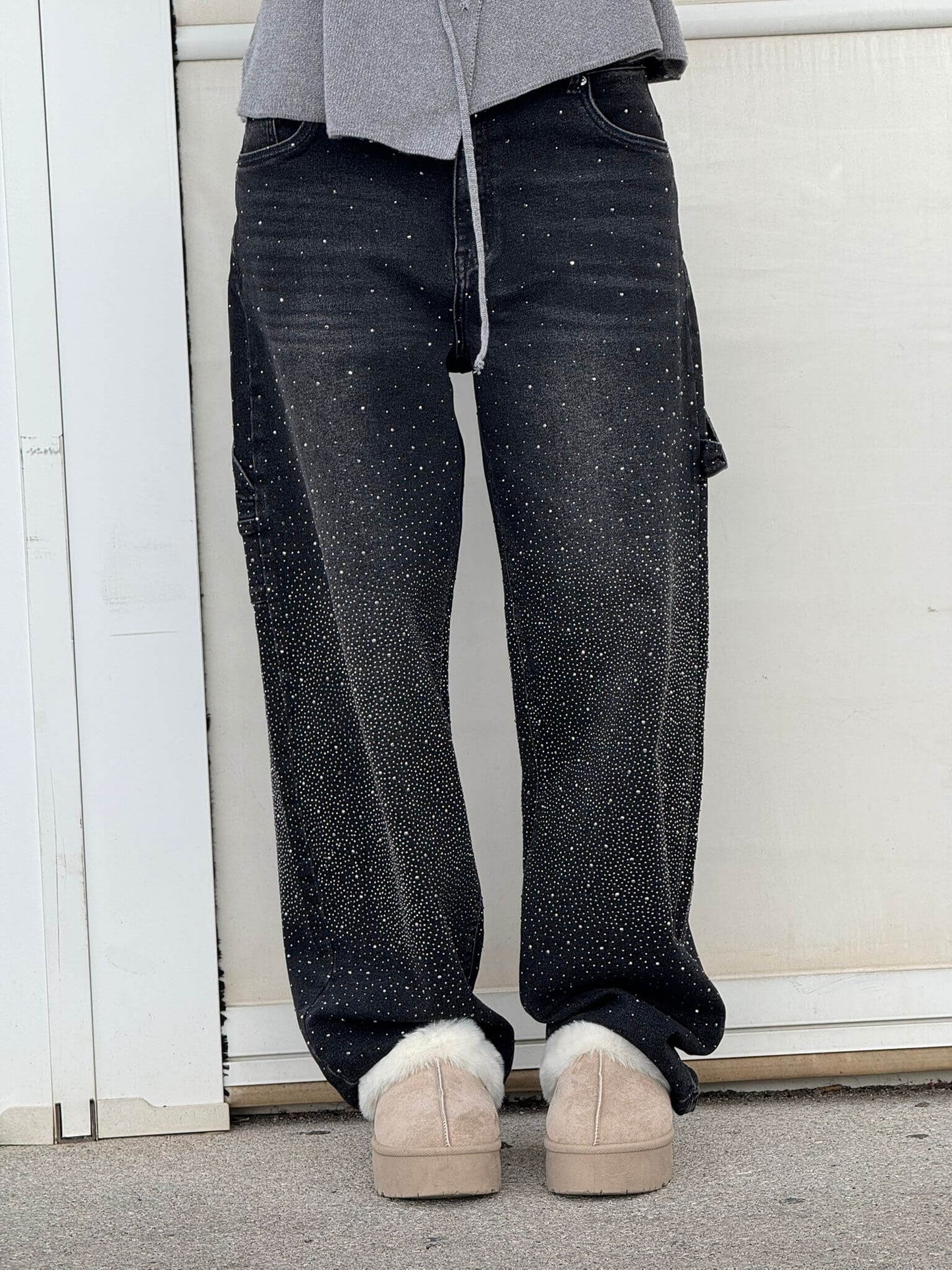Person wearing black speckled jeans and beige shoes against a white wall.