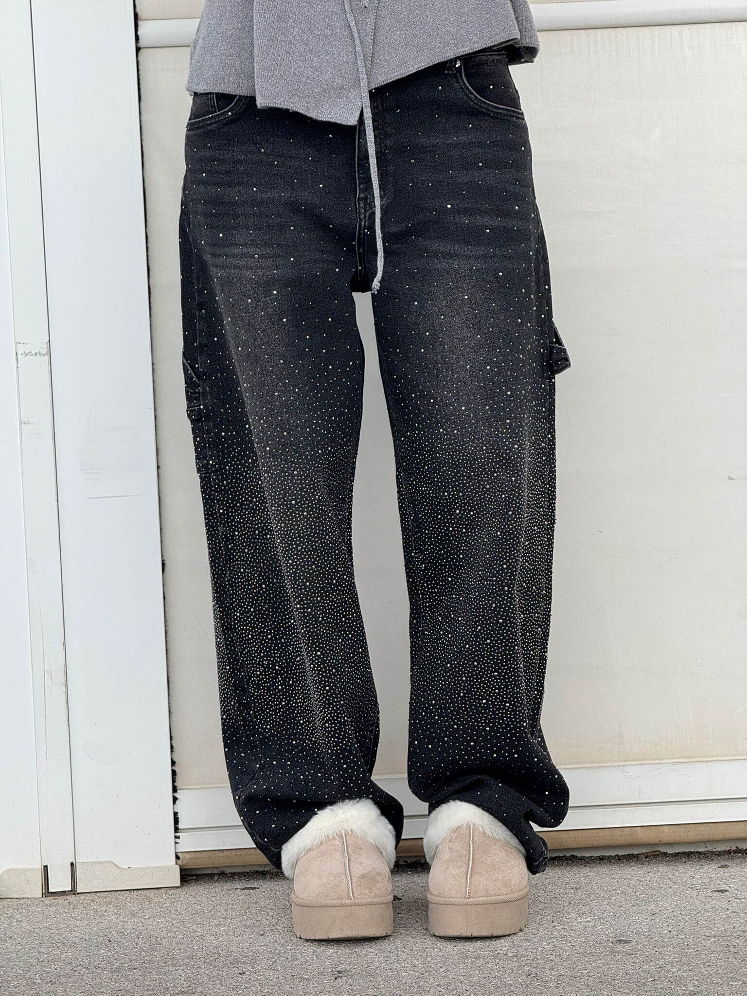 Person wearing black speckled jeans and beige shoes against a white wall.