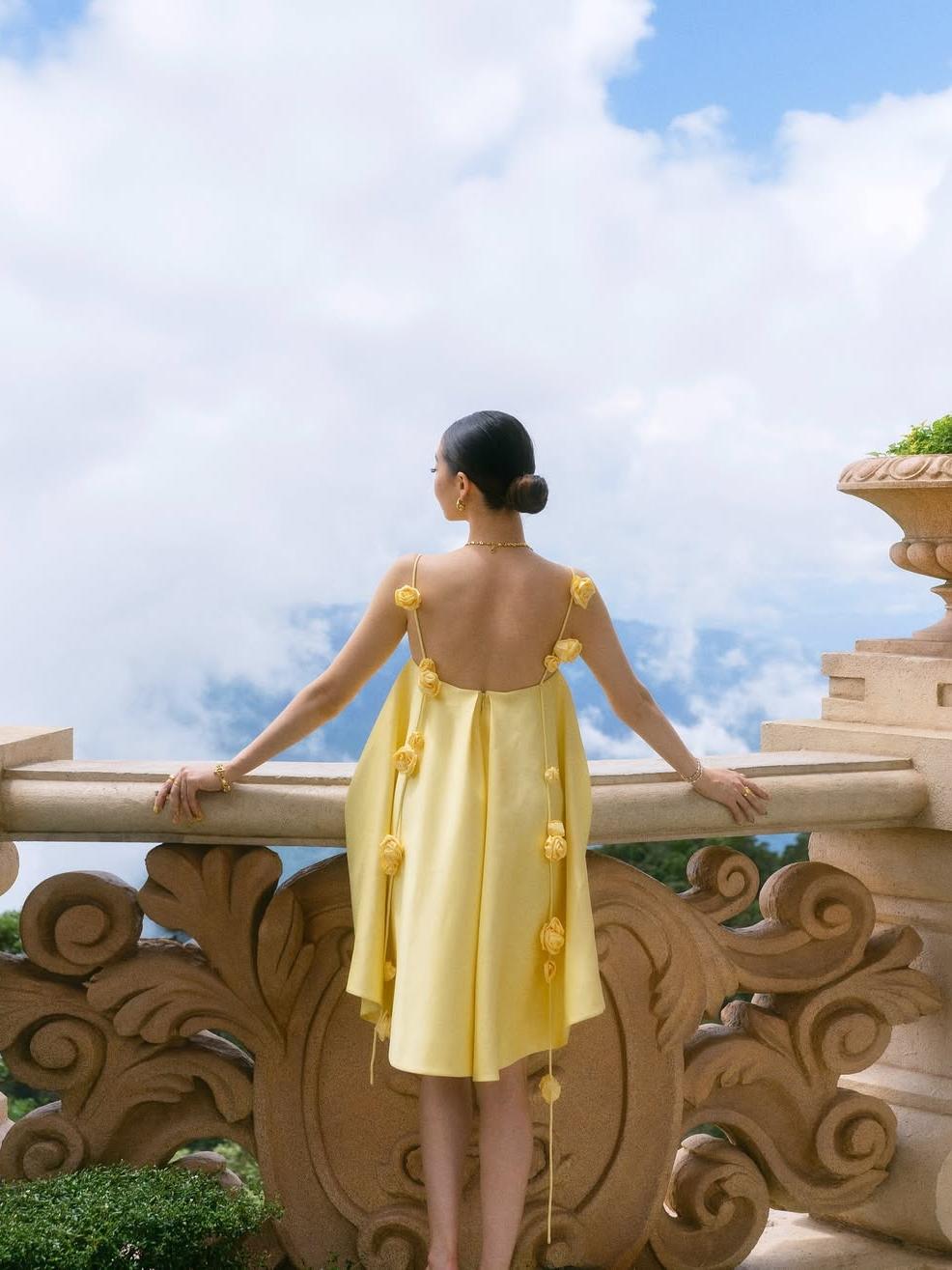Woman in a yellow dress standing on a balcony with a blue sky and clouds in the background